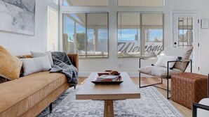 Un salon moderne avec un canapé, un fauteuil et une table basse. De grandes fenêtres laissent entrer la lumière naturelle et offrent une vue sur l’extérieur.