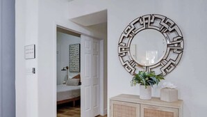 Un couloir avec un miroir rond, une vitrine contenant des objets décoratifs et une porte donnant sur une chambre à coucher.