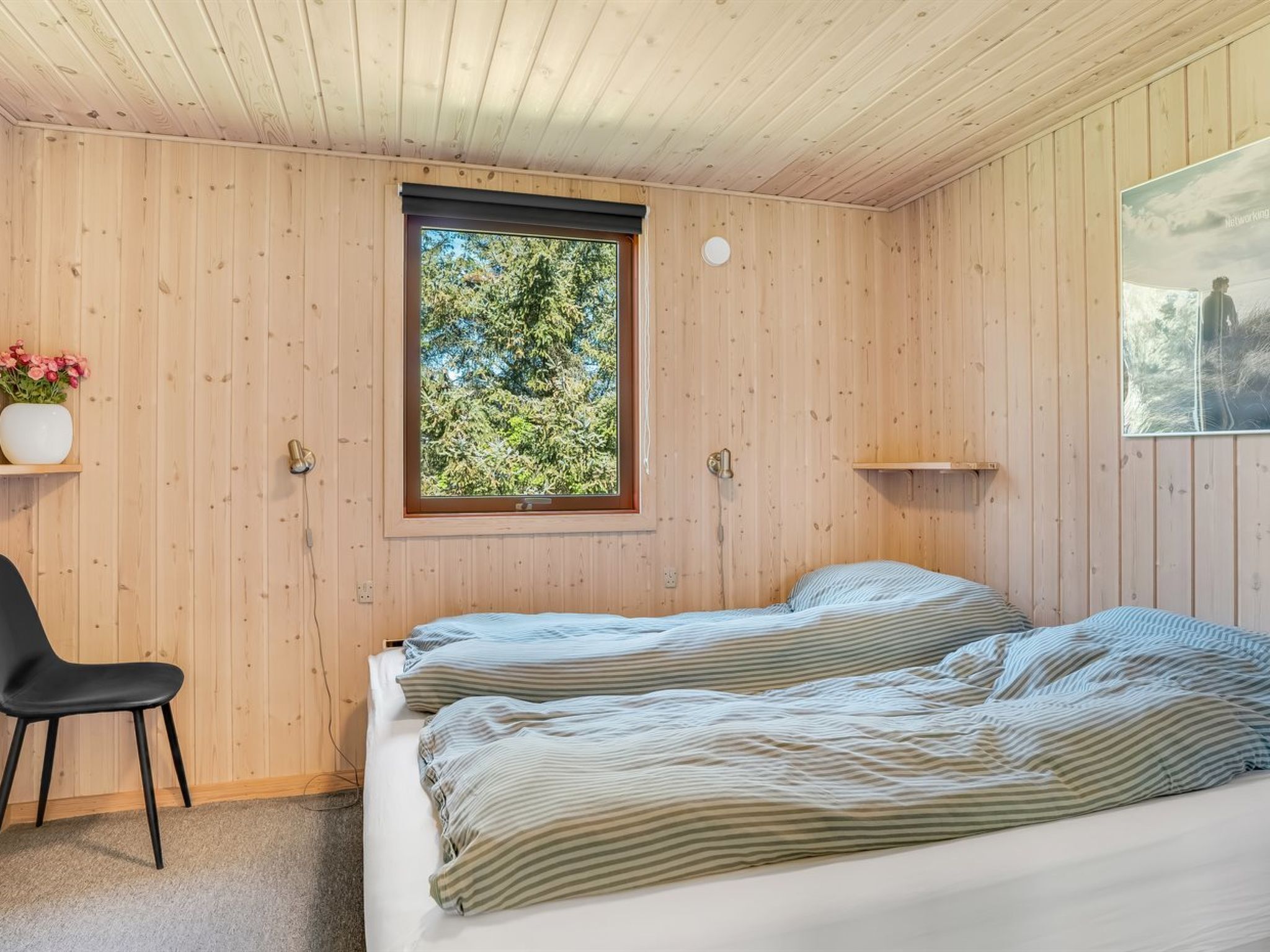 A bedroom with wooden walls, a window with a view of trees, a bed with striped bedding, a chair, and a small shelf.