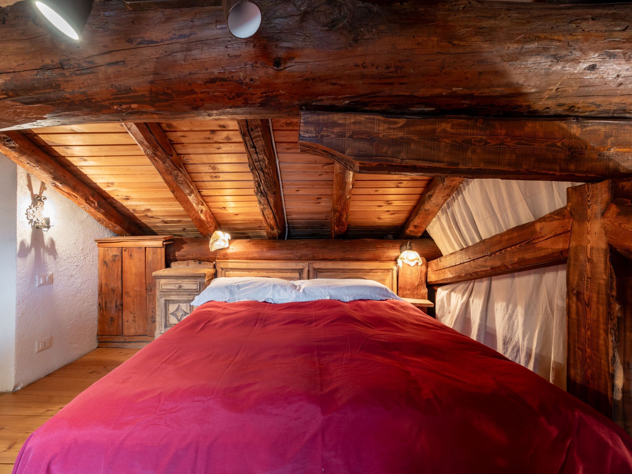 A cozy cabin-style bedroom with a wooden ceiling, a bed with a red blanket, and two wall-mounted lamps.