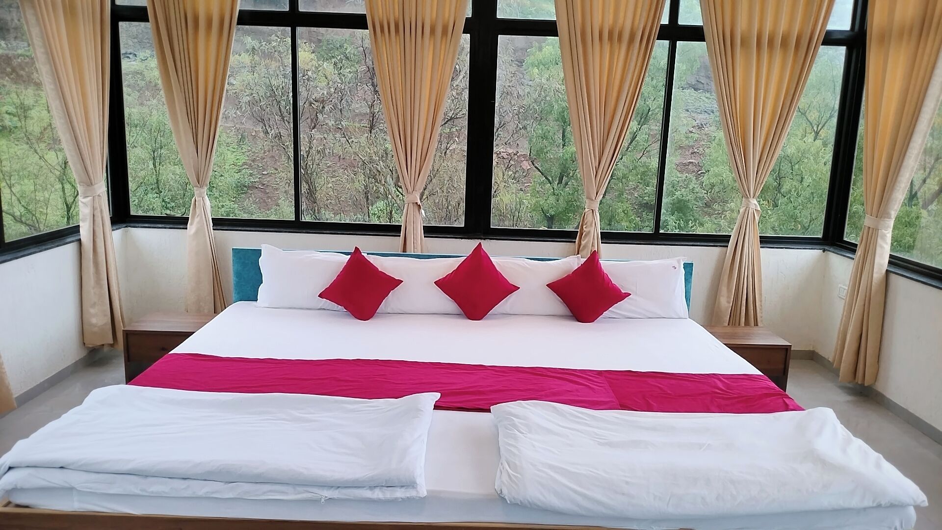 A bed with white and red bedding, a view of trees through large windows, and beige curtains.