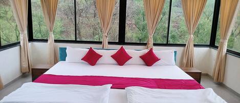 A bed with white and red bedding, a view of trees through large windows, and beige curtains.