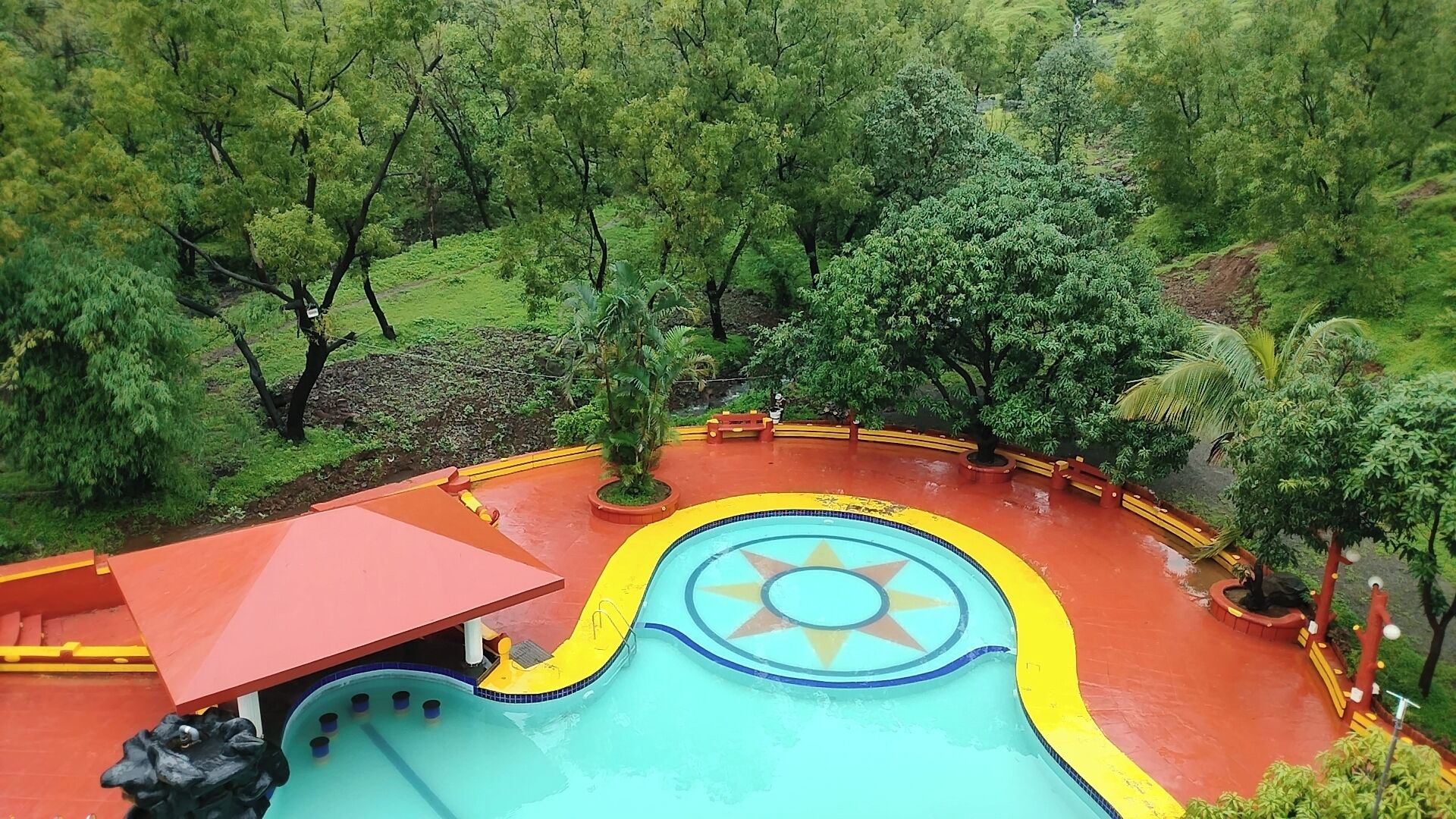A swimming pool with a star-shaped design, surrounded by a red and yellow pathway, set in a lush green forest.