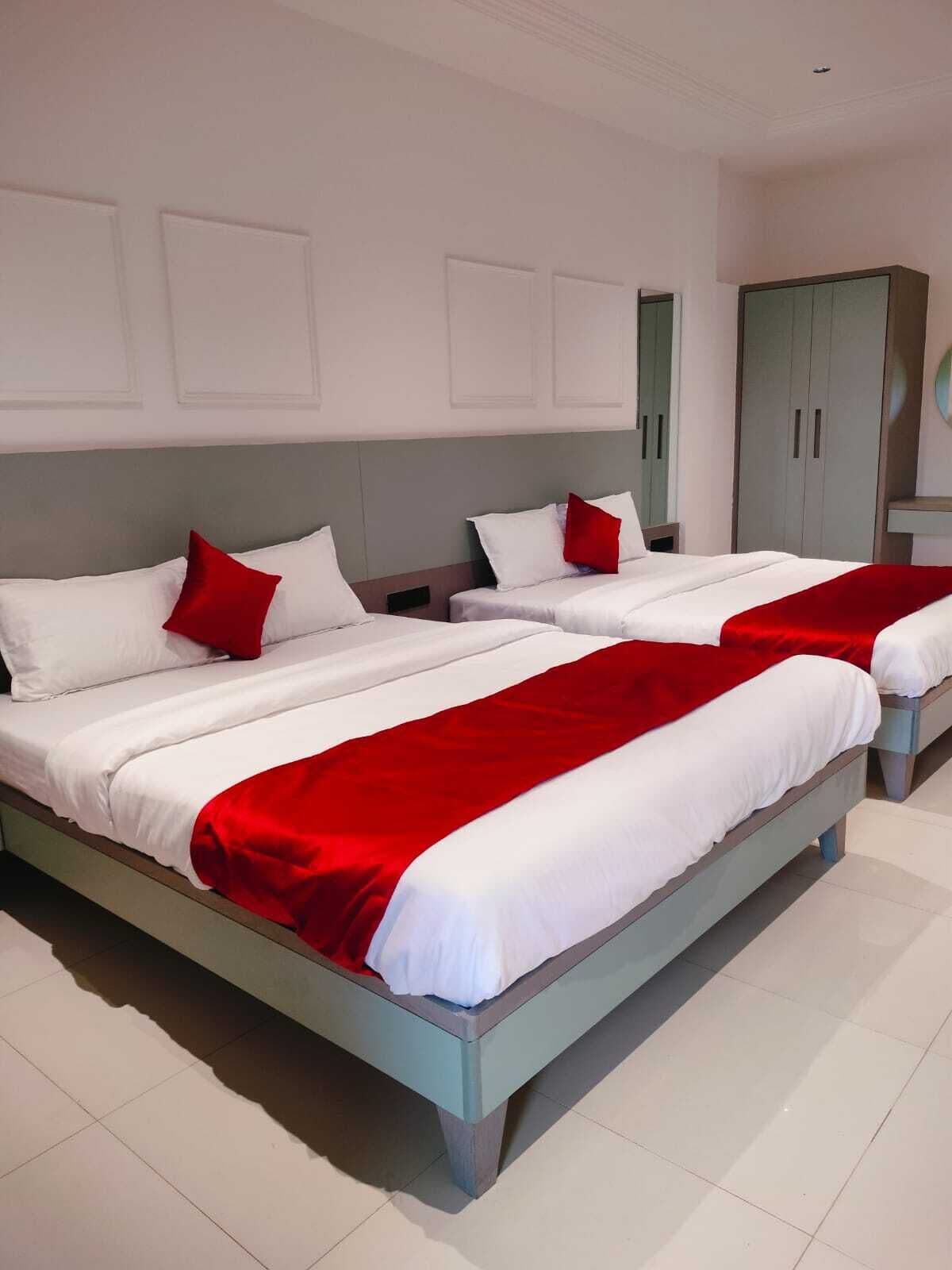 A hotel room with two beds, white bedding, red accents, a grey wardrobe, and a white tiled floor.