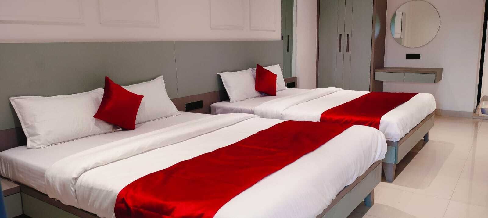 A hotel room with two beds, each with white bedding and red accents, a bedside table, a mirror, and a wardrobe.