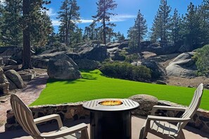 A patio with a fire pit, two Adirondack chairs, and a view of a green lawn surrounded by rocks and trees.