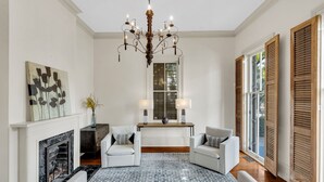 Una sala de estar con chimenea, dos sillones blancos, un sofá, una araña y un ventanal con contraventanas.
