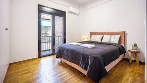 A bedroom with a wooden floor, a bed with a dark-colored duvet, a wooden headboard, a small bedside table with a lamp, and a door leading to a balcony. - 2BRCozy Urban Retreat Exarchia (Athens)