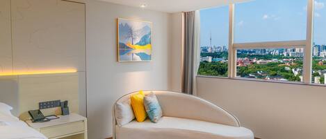 Habitación de hotel moderna con sofá curvo, mesita de noche con teléfono y cargador, un cuadro en la pared y un ventanal con vistas a la ciudad.