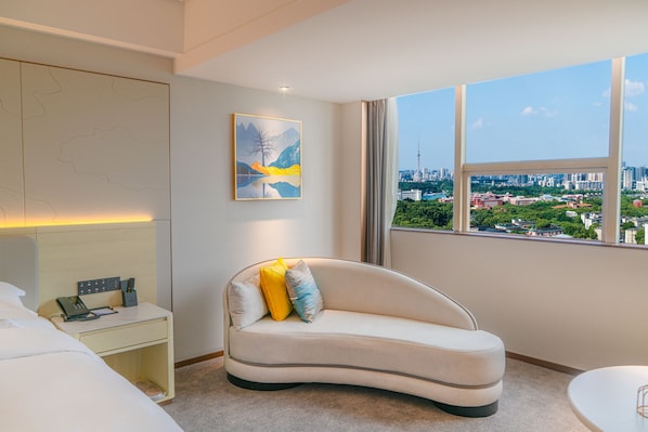 A modern hotel room with a curved sofa, a bedside table with a phone and charger, a painting on the wall, and a large window offering a city view. - Li Gao Exclusive Hotel (Changsha)