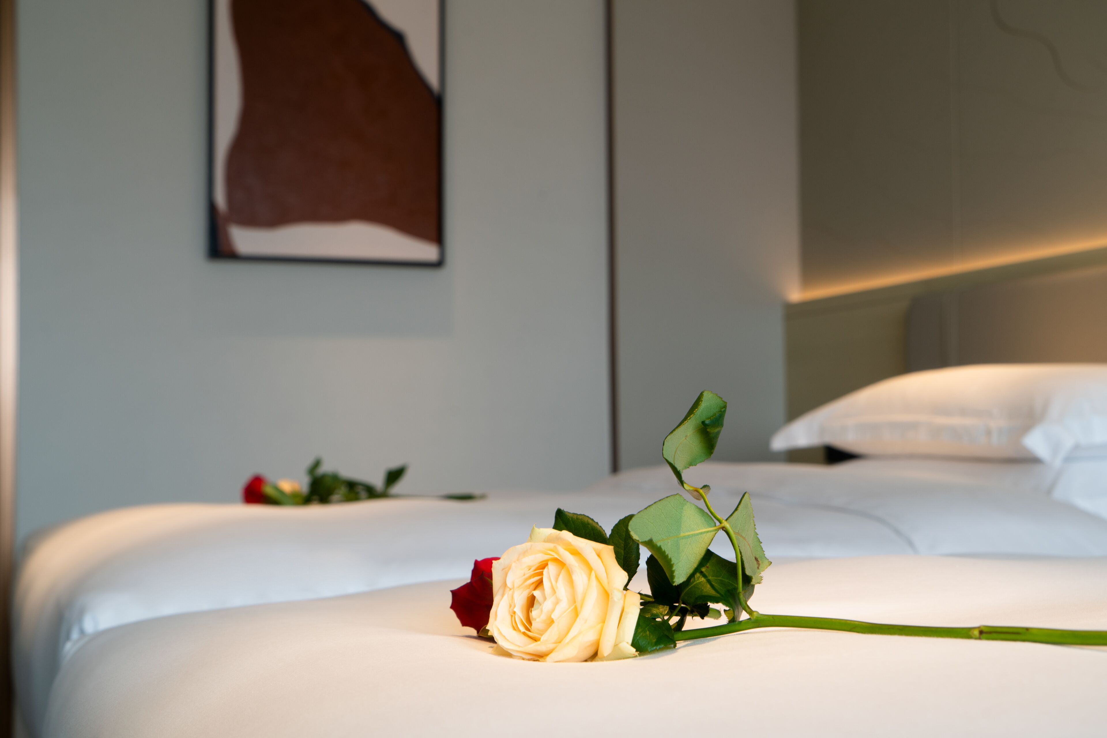 Una habitación de hotel con una cama bien tendida, una rosa sobre la cama y un cuadro en la pared.