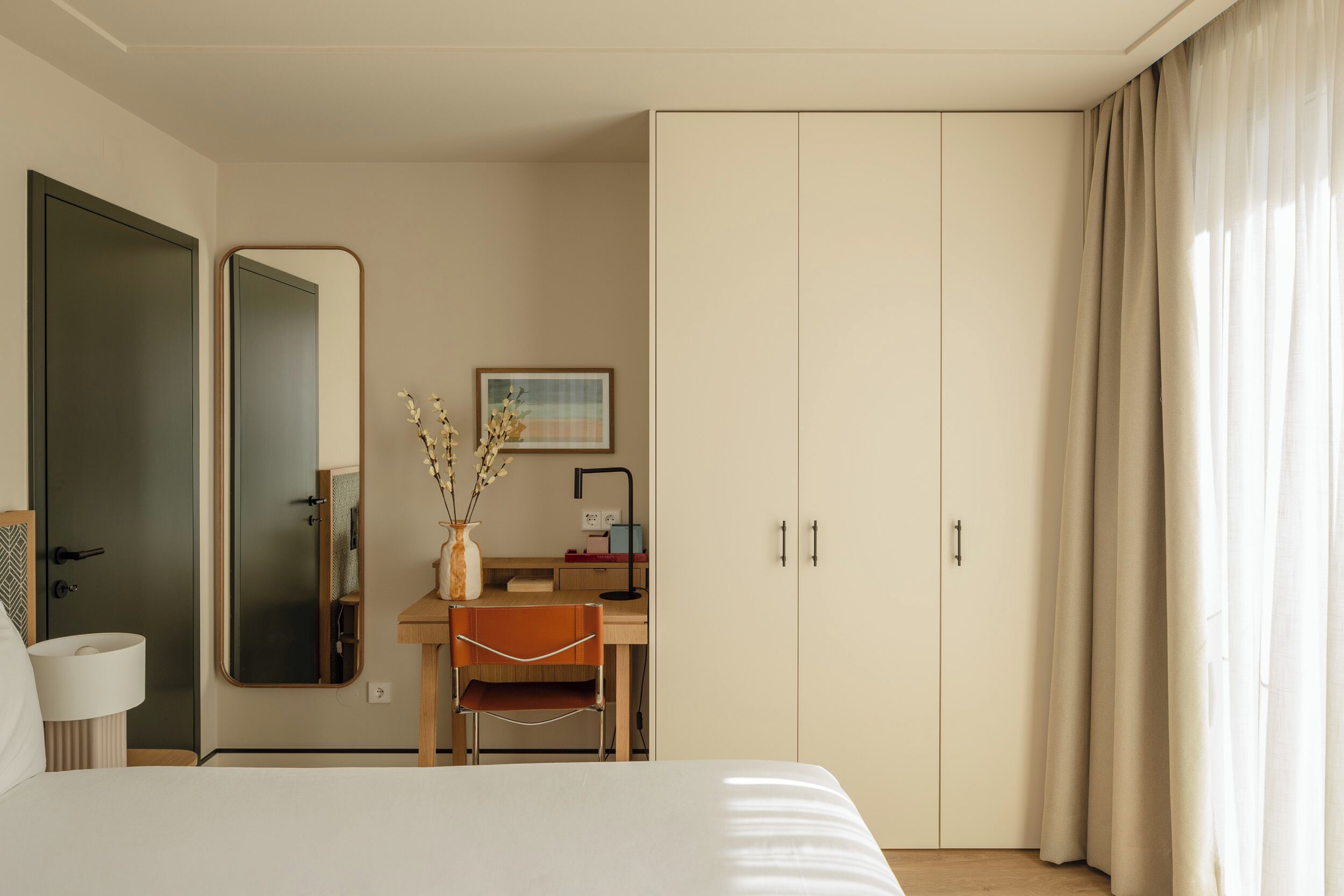 A hotel room with a bed, a desk with a chair, a wardrobe, a mirror, and a vase with flowers.