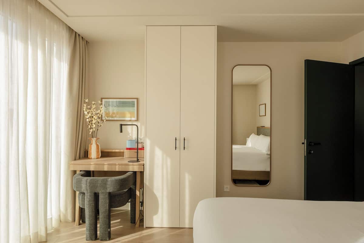 A hotel room with a bed, a desk with a chair, a wardrobe, a mirror, and a window with curtains.