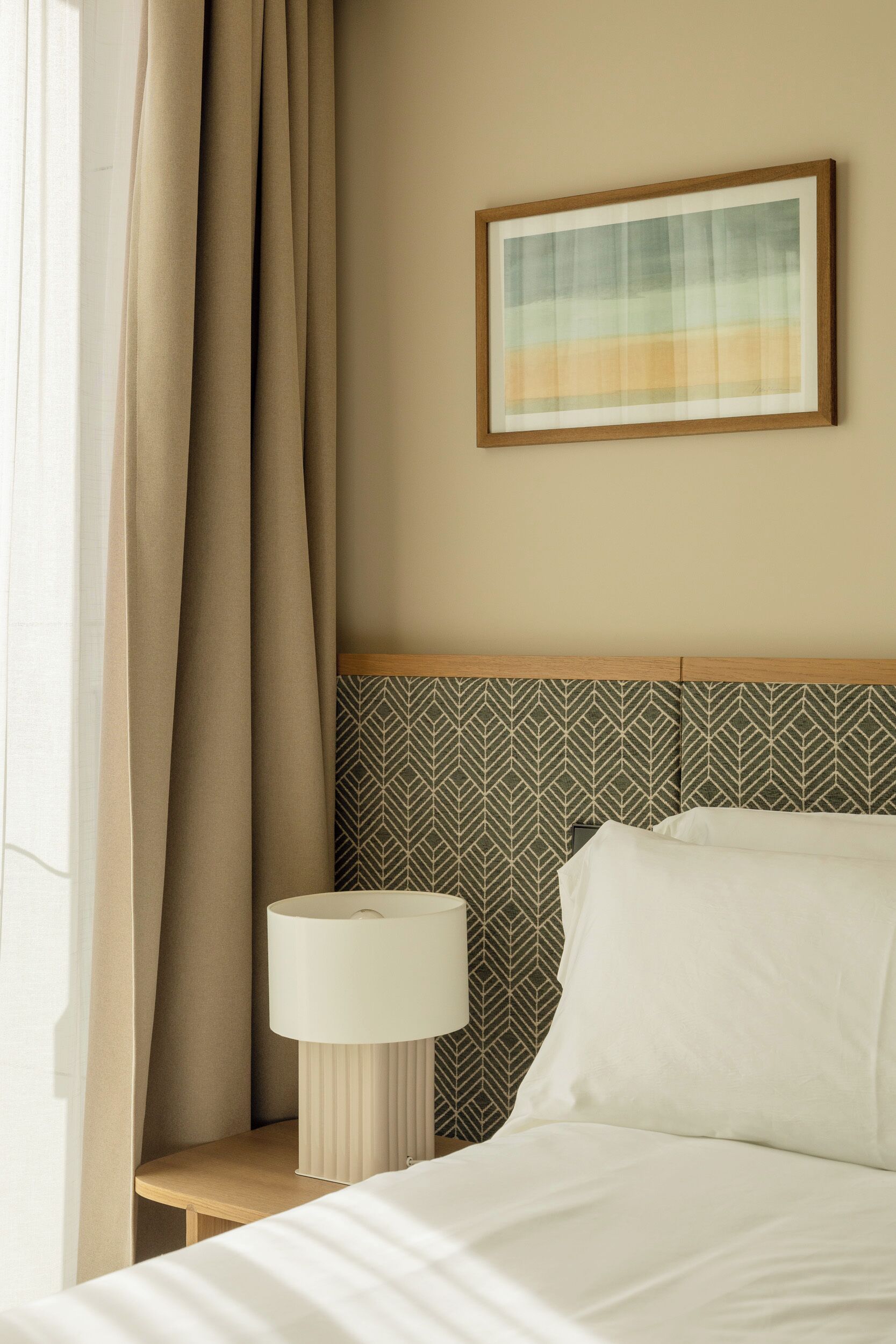Een net opgemaakt bed met een hoofdbord met een geometrisch patroon, een bedlampje en een ingelijst foto aan de muur.