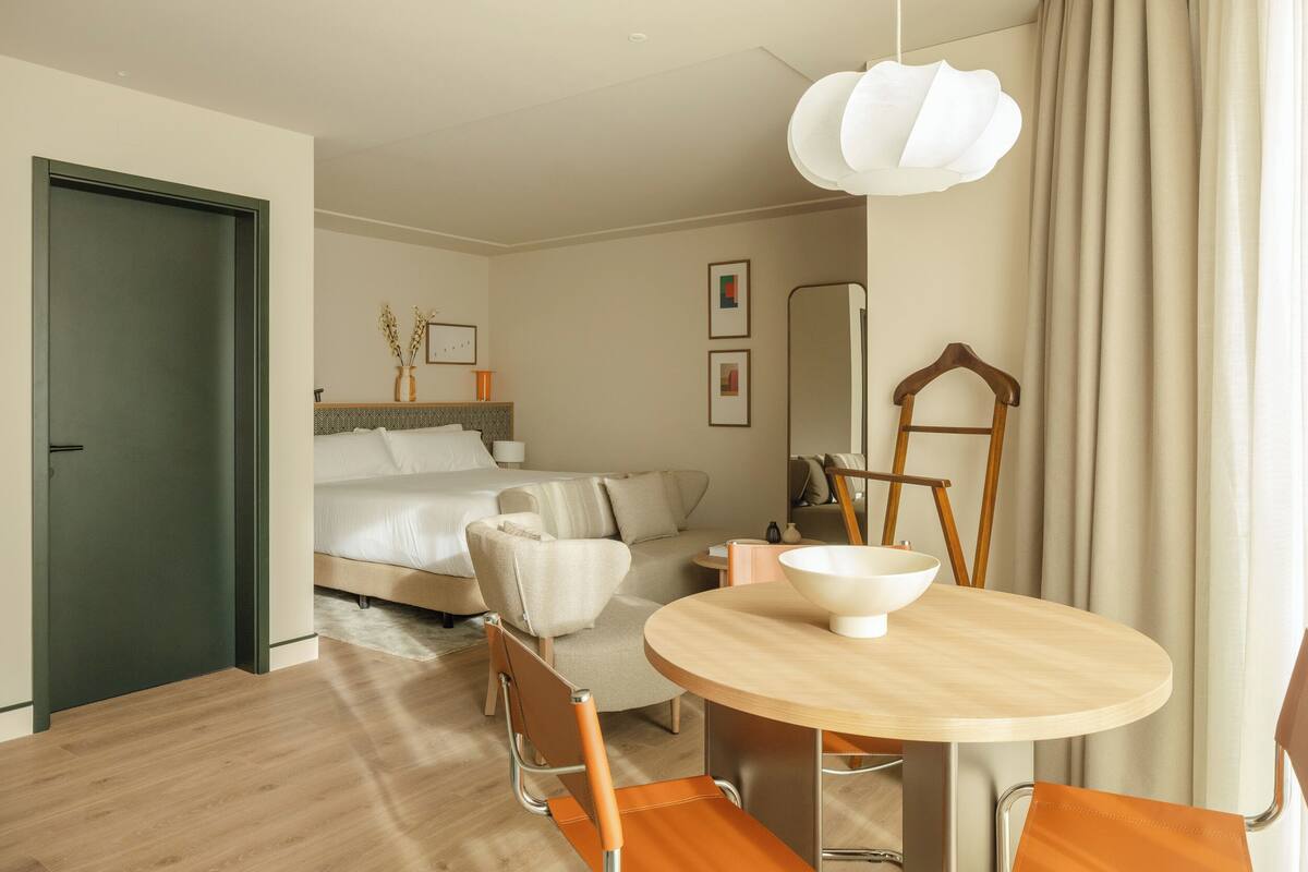 A modern hotel room with a bed, a sitting area with a sofa and chairs, a dining table with a bowl, and a mirror.