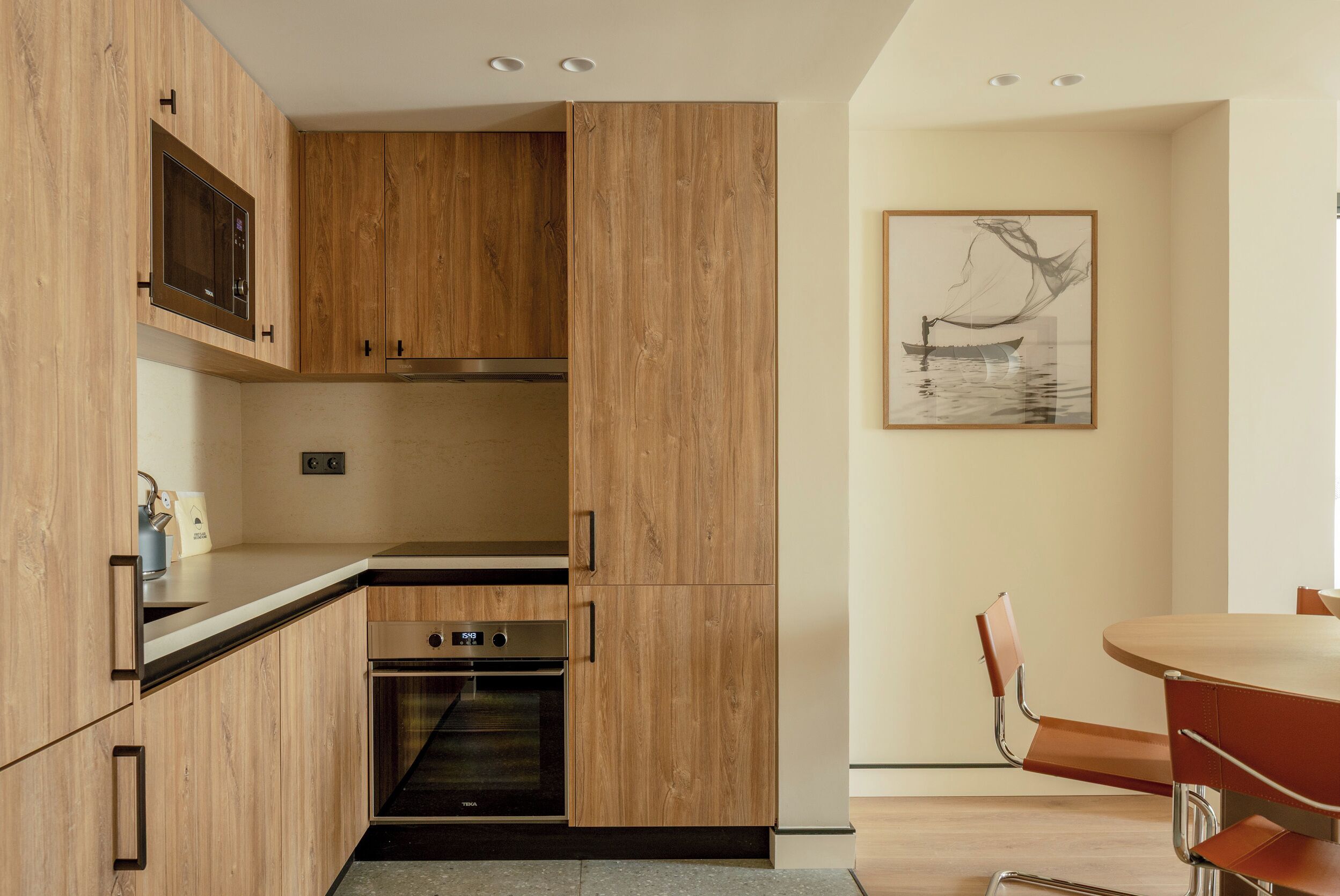 A modern kitchen with wooden cabinets, a built-in microwave, and an oven. There is a dining area with a table and chairs.