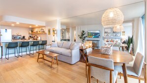 Ein modernes Wohnzimmer mit Sofa, Couchtisch und Essbereich. Im Hintergrund ist eine Küche mit Barhocker zu sehen.