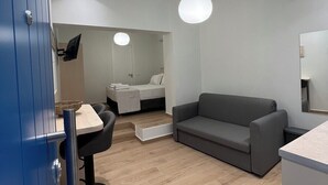 A modern, well-lit room with a grey sofa, a small dining table with chairs, a bed with a white headboard, and a large mirror on the wall. - Cozy appartment at the heart of the Historic center - Hermes Central Suite (Αθήνα)