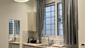 A modern kitchen with a built-in dishwasher, a coffee machine, and a sink with a faucet. There is a window with curtains, a ceiling light, and a fire extinguisher on the wall. - Cozy appartment at the heart of the Historic center - Hermes Central Suite (Αθήνα)