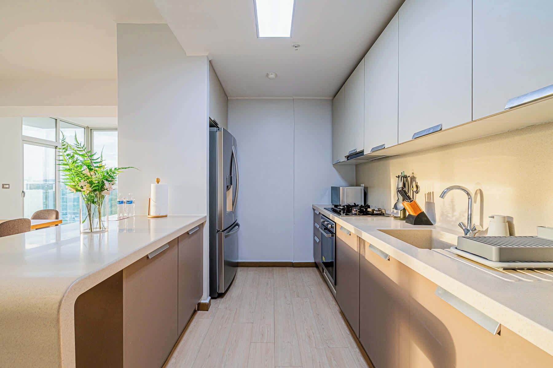 Une cuisine moderne équipée d’un réfrigérateur encastré, d’une cuisinière et d’un évier. Une fenêtre laisse entrer la lumière naturelle et offre une vue sur la ville.