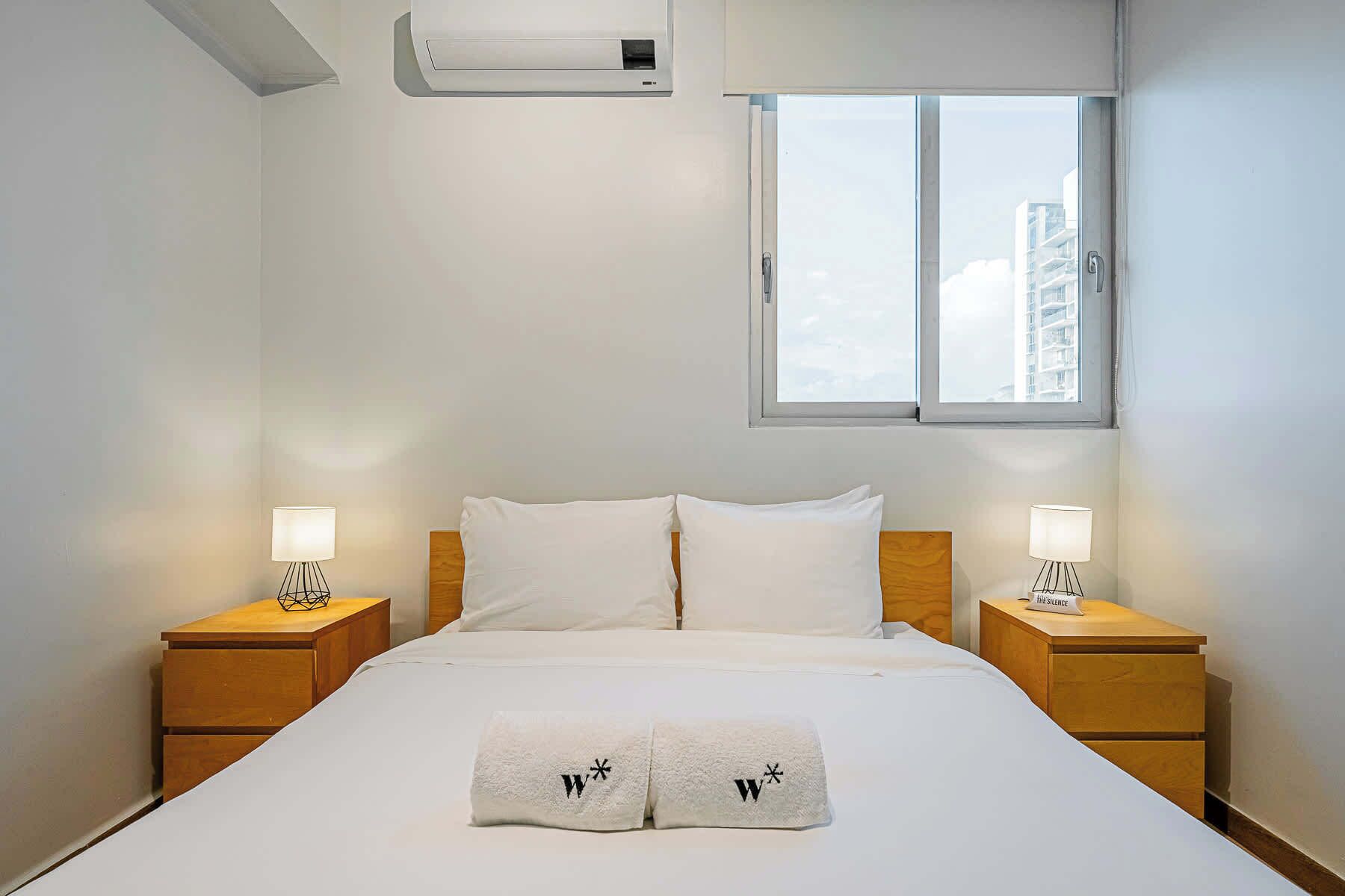 Habitación de hotel con una cama grande, dos mesitas de noche con lámparas, una ventana con vistas a edificios y un equipo de aire acondicionado sobre la cama.