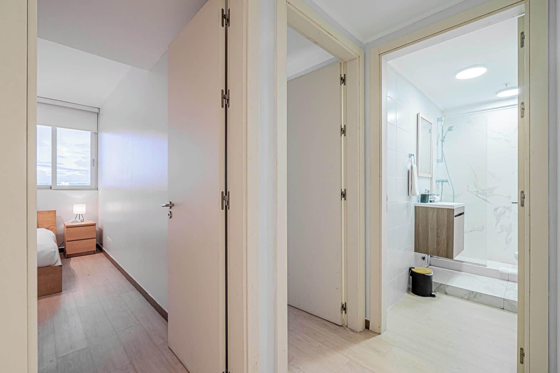 A modern hotel room with a wooden bed, a bedside table, a nightstand, a window, and a bathroom with a shower, mirror, and sink.
