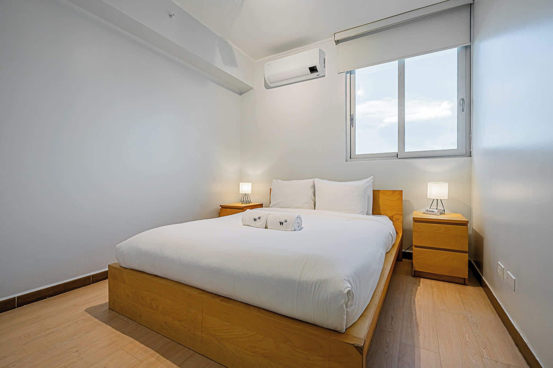 A modern hotel room with a large bed, two bedside tables with lamps, a window with a view, and an air conditioning unit on the wall.