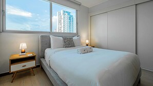 A modern hotel room with a large bed, a nightstand with a lamp, a window with a city view, and a wardrobe. - w* | Airy 2BR in San Francisco w/ Pool (Panamá)