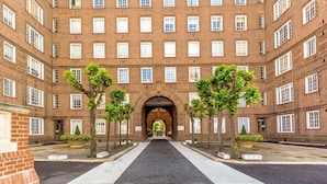 Un immeuble en briques à plusieurs étages, doté d’une arche centrale et d’une allée bordée d’arbres disposés de manière symétrique.