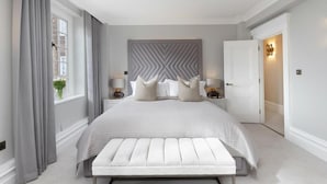 Une chambre à coucher avec un grand lit, une tête de lit ornée d’un motif géométrique, deux tables de chevet avec des lampes, une fenêtre avec des rideaux et une porte avec un luminaire.