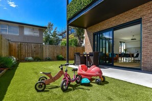 Een achtertuin met kunstgras, een houten schutting en een huis van baksteen met een schuifdeur die uitkomt op een eethoek.