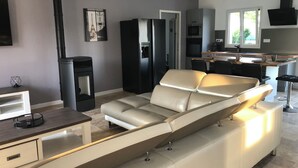 Uma sala de estar moderna com sofá, lareira, área de cozinha com mesa de jantar e uma grande janela que proporciona iluminação natural.