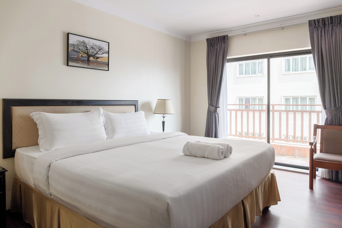 A hotel room with a large bed, a nightstand, a lamp, a chair, and a balcony with curtains.