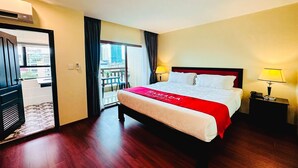 Une chambre d’hôtel avec un grand lit, une couvre-lit rouge, deux lampes de chevet, une table de chevet et une vue sur la ville depuis la porte d’un balcon.