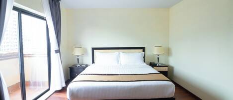 A hotel room with a large bed, two bedside tables with lamps, a window with curtains, and a view of buildings outside.