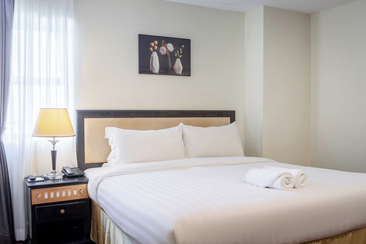 A hotel room with a bed, a nightstand with a lamp, a painting on the wall, and a window with curtains.