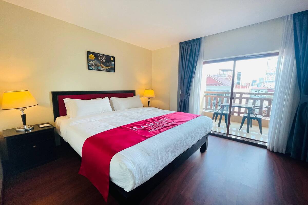 Une chambre d’hôtel avec un grand lit, deux lampes de chevet, un plaid rouge, un tableau au mur et un balcon aménagé avec du mobilier d’extérieur.