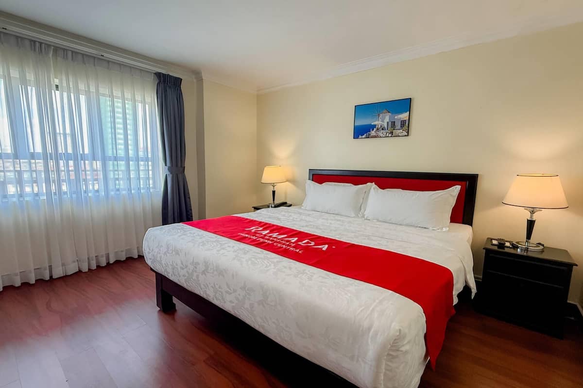 Une chambre d’hôtel avec un grand lit, deux tables de chevet avec des lampes, une fenêtre avec des rideaux légers et un tableau au mur.
