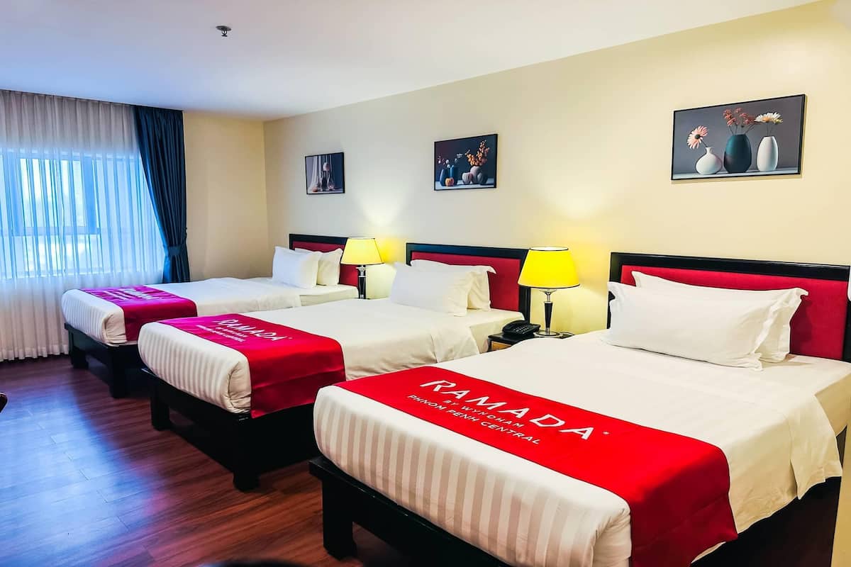 Une chambre d’hôtel avec trois lits, chacun recouvert d’une couvre-lit rouge et blanche, et dotée d’une tête de lit rouge. Il y a deux tables de chevet avec des lampes, et la chambre dispose d’une grande fenêtre avec des rideaux.