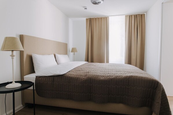Hotellihuone, jossa on sänky, kaksi yöpöytälamppua, ikkuna verhoilla ja pieni pöytä.