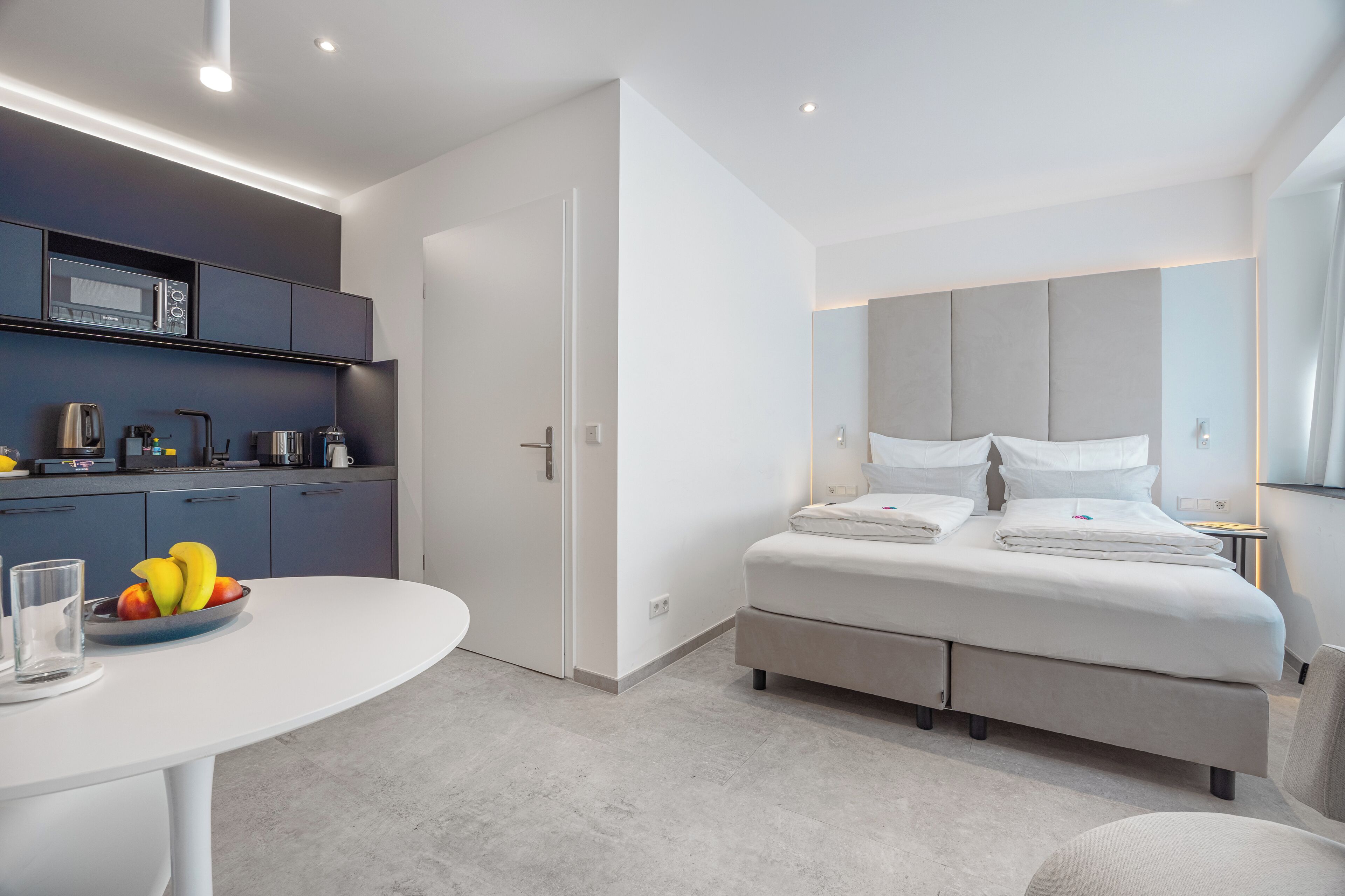 A modern hotel room with a bed, bedside table, and a kitchenette area.
