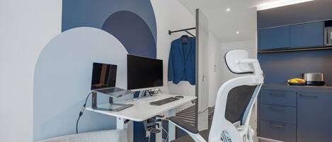 Oficina moderna con escritorio, computadora y silla ergonómica. Se observa un perchero con una chaqueta azul y una zona de cocina con armarios y microondas.