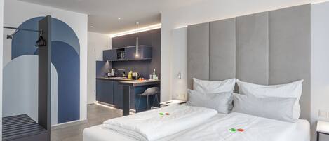 A modern hotel room with a large bed, a desk, and a kitchenette.