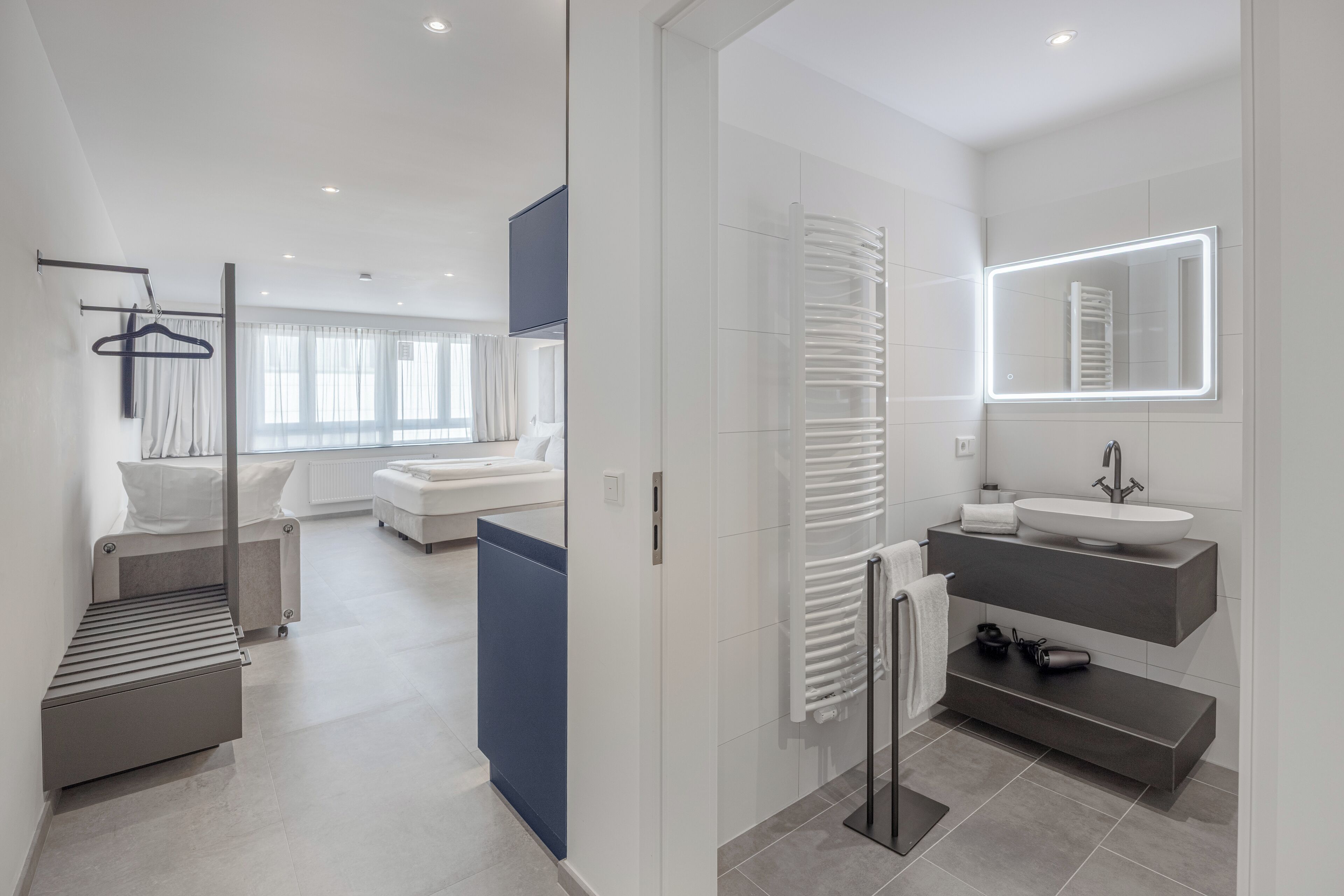 A modern hotel room with a large bed, a separate sitting area, a bathroom with a vanity and a mirror, and a view of the city.
