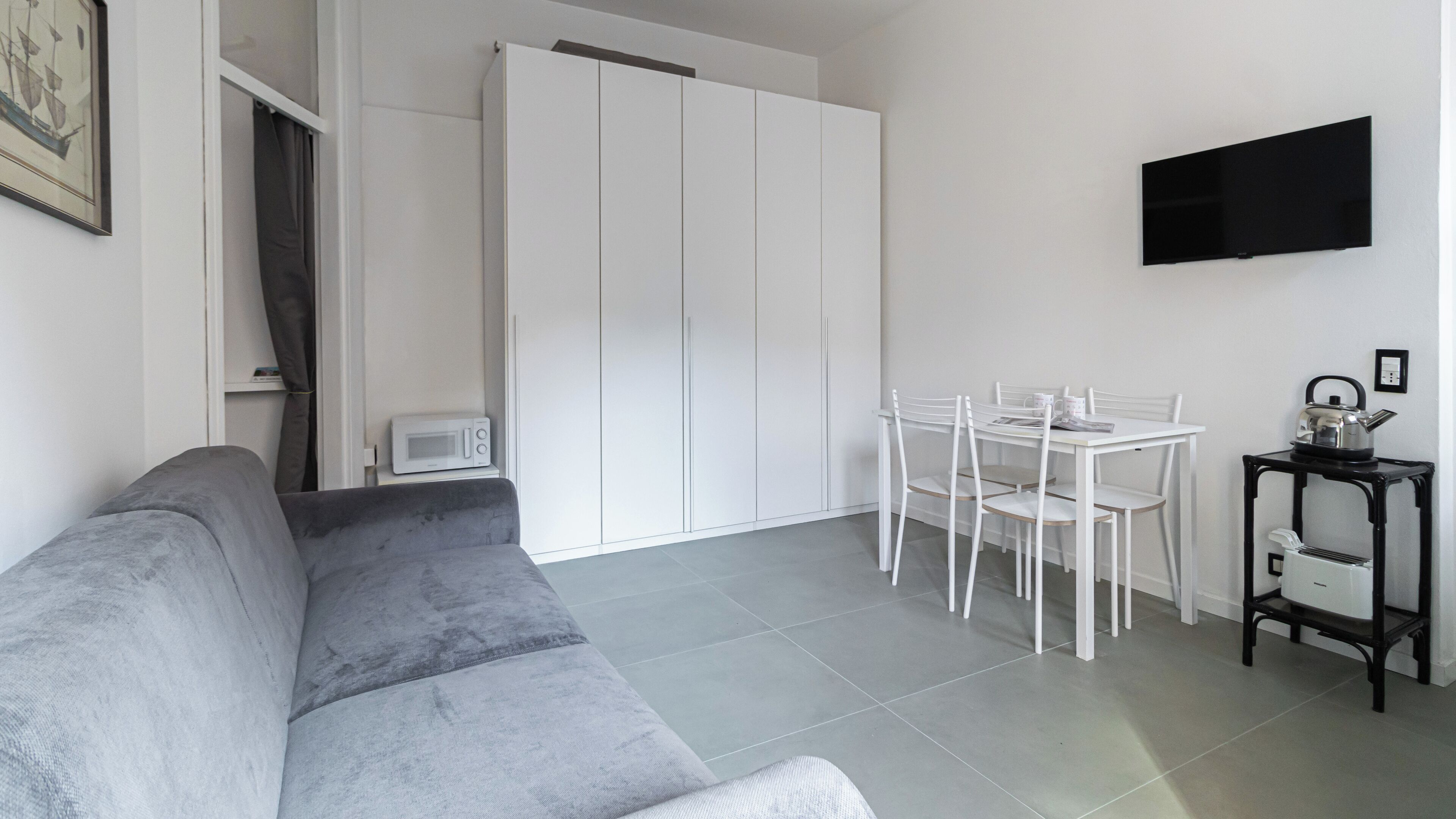 A modern living room with a grey sofa, a dining table with chairs, a white wardrobe, a microwave, a kettle, and a TV on the wall.