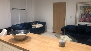 Uma sala de estar moderna com um sofá azul escuro, uma poltrona branca e uma mesa de centro de madeira. Há um quadro na parede e uma porta visível ao fundo.