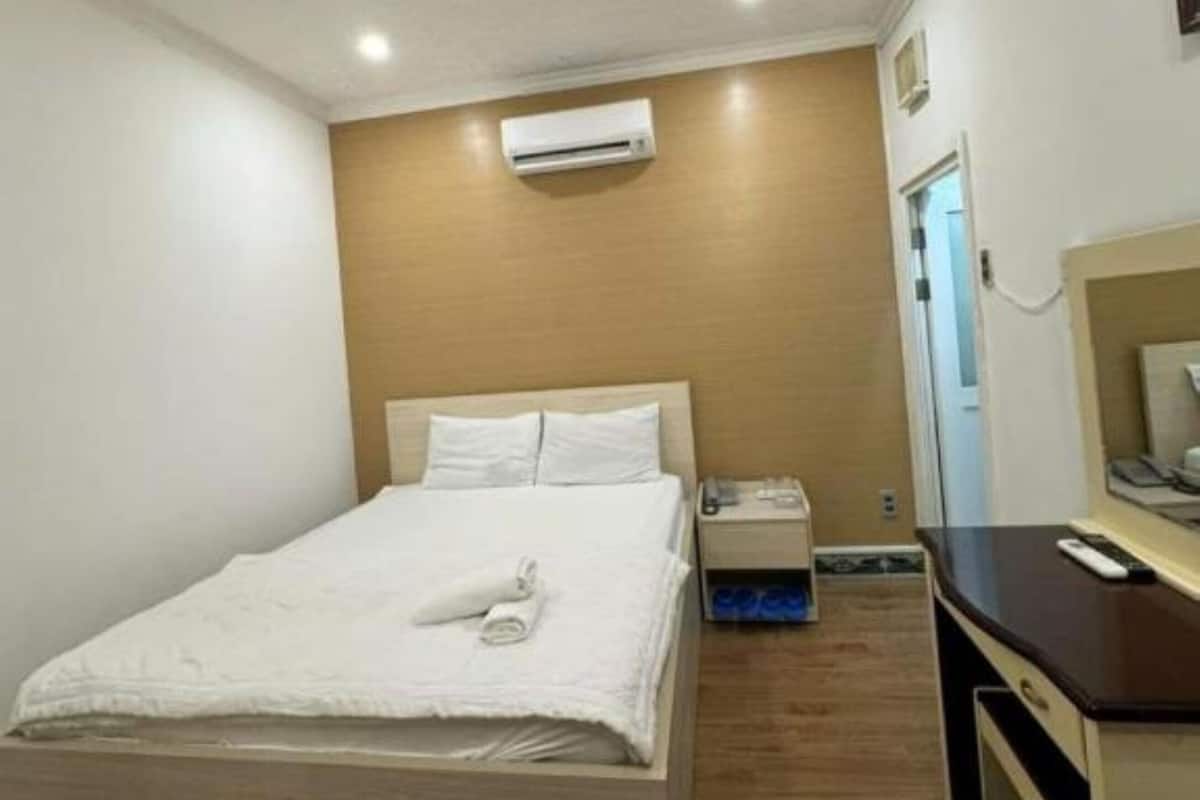 Une chambre d’hôtel avec un plancher en bois, un lit avec du linge de maison blanc, une table de chevet avec une lampe et un appareil de climatisation fixé au mur.