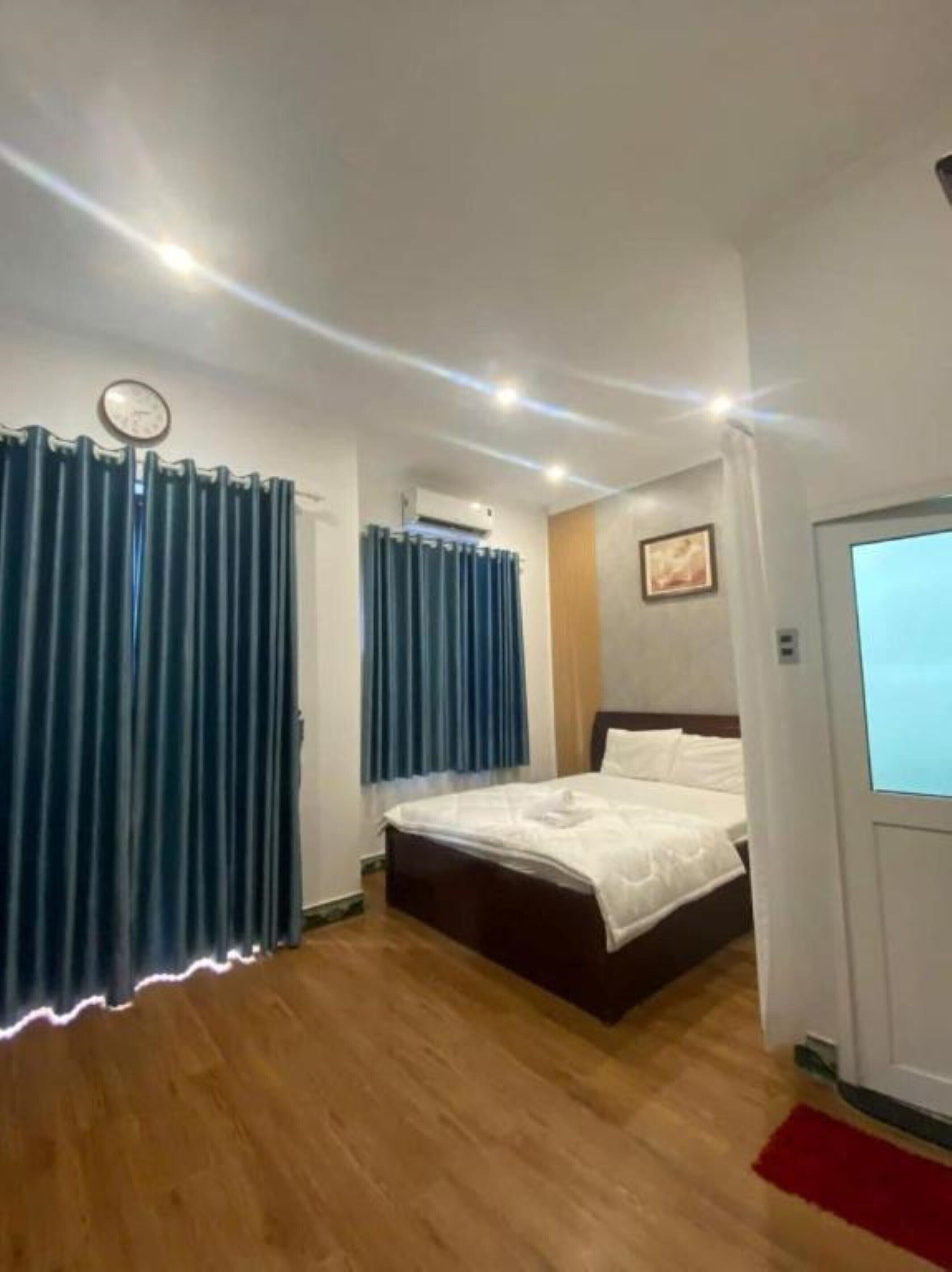 Une chambre d’hôtel avec un plancher en bois, un lit recouvert de draps blancs, une horloge au mur et des rideaux bleus.