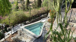 Une piscine rectangulaire bordée de pierres, entourée d’un chemin également en pierres et de différentes plantes.