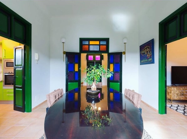 Un comedor con una mesa central, sillas, una planta en maceta y una pared de colores con un diseño geométrico.